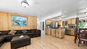 A spacious living room with a large sectional sofa, a wooden dining table, and a kitchen area with wooden cabinets and stainless steel appliances. - Moonlight Serenity (Big Bear Lake)