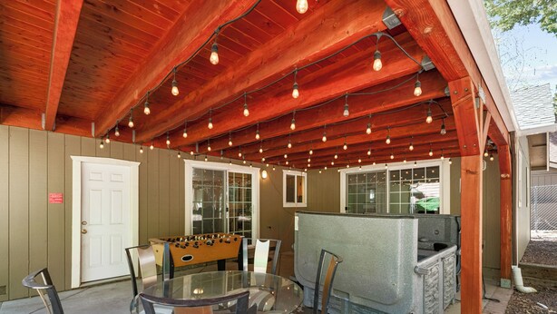 An outdoor patio with a round glass table and chairs, a foosball table, and a covered area with wooden beams and string lights. - Moonlight Serenity (Big Bear Lake)