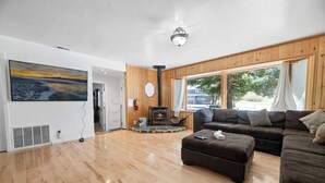 A spacious living room with a wooden floor, a large sofa set, a fireplace, and a large window offering a view of the outdoors. - Moonlight Serenity (Big Bear Lake)