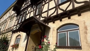 Un edificio tradicional de entramado de madera con una entrada destacada, un pequeño jardín con flores rojas y un cielo azul despejado.