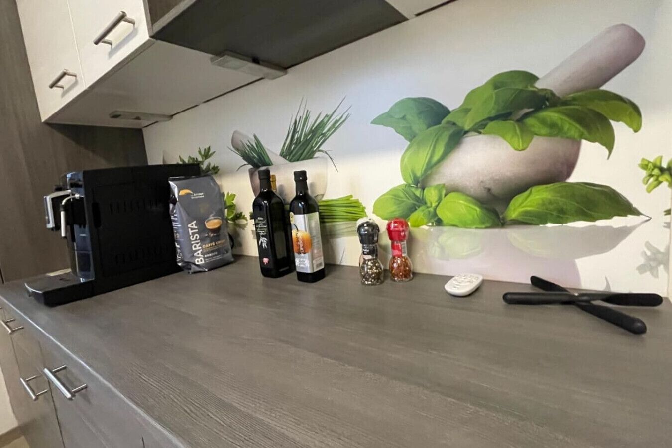 A kitchen counter with a coffee machine, a bag of coffee, bottles of olive oil, salt and pepper shakers, a remote control, and a knife.