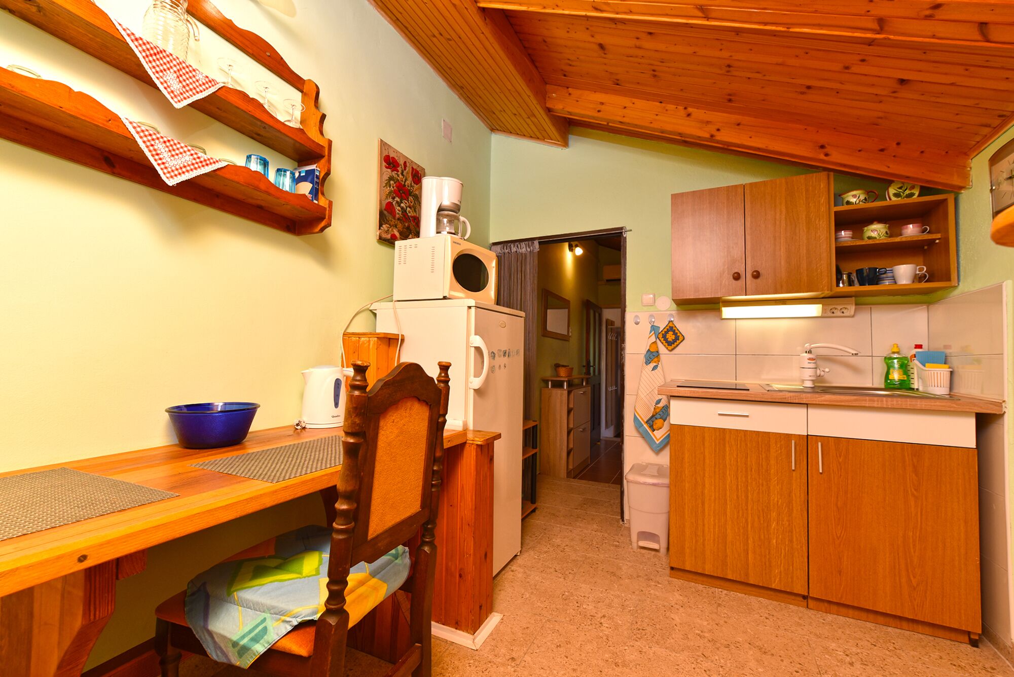 A compact kitchen with wooden cabinets, a refrigerator, a microwave, and a sink. There is a dining table with chairs and a blue bowl on it.