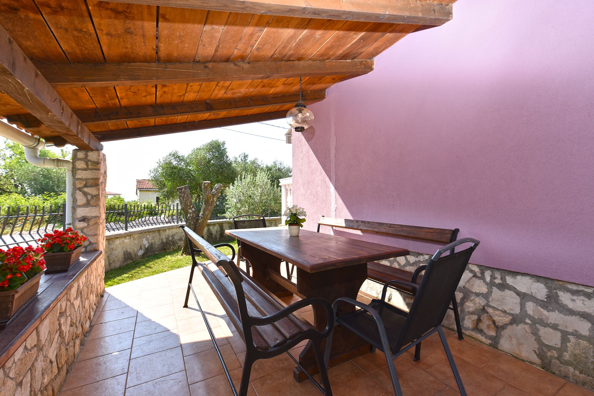 Une terrasse extérieure avec une table et des chaises en bois, un mur en pierre et une vue sur la verdure et les bâtiments en arrière-plan.