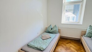 A small, bright room with a wooden herringbone floor, a single bed with a green patterned mattress and pillows, and a window with blinds. - Leoben Apartment Top 6 (Leitendorf)