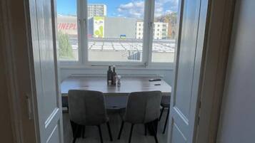 Kapea käytävä, jonka päässä on ruokailutila, jossa on pöytä ja tuolit. Ikkunasta avautuu näkymä rakennuksiin ja selkeälle taivaalle.