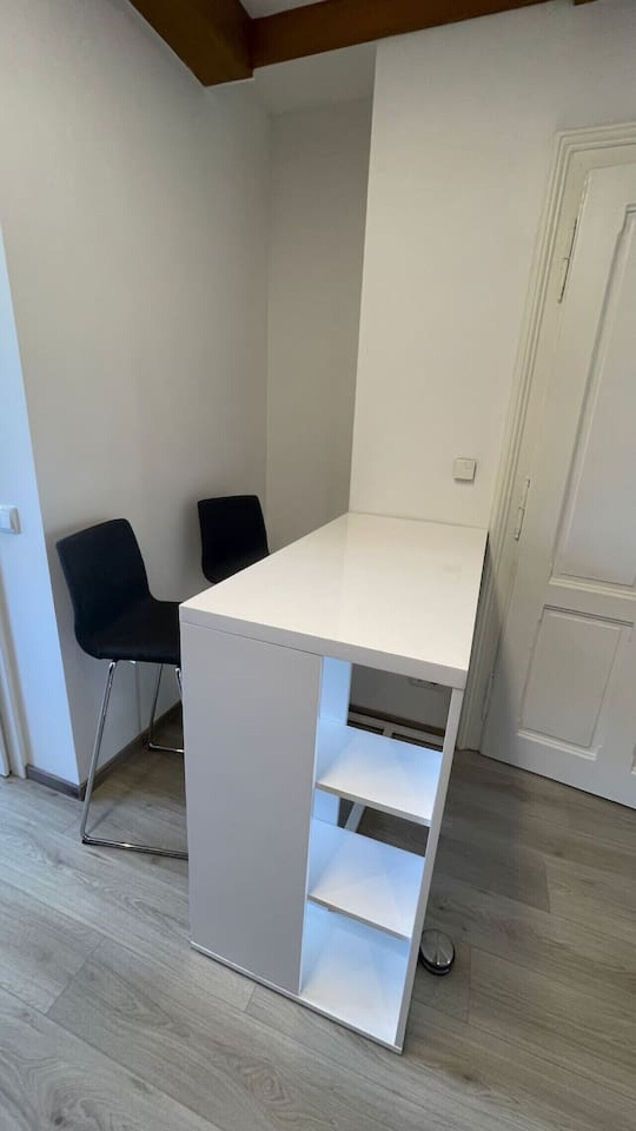 A small, white, L-shaped desk with two shelves, black chairs, and a white door in a room with light wooden flooring.