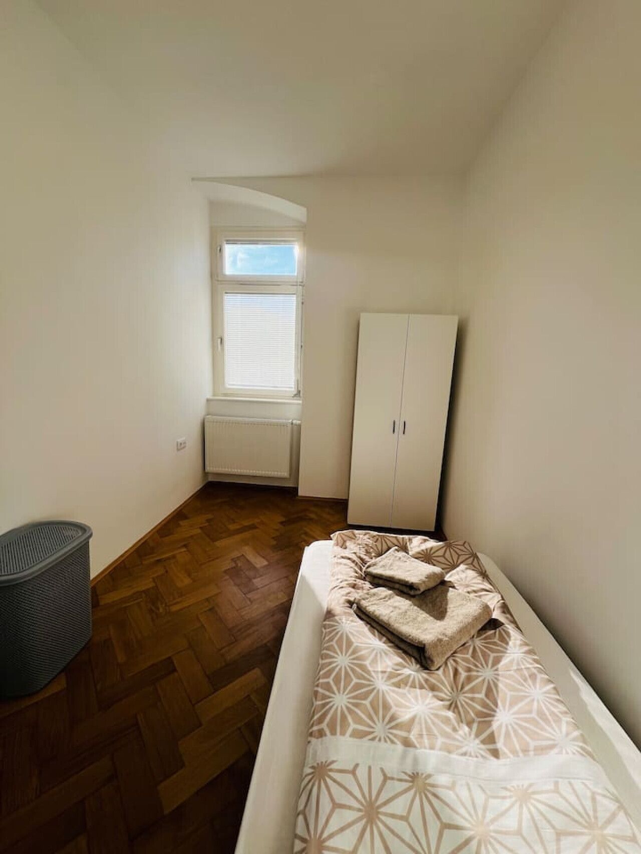 Una habitación pequeña y poco amueblada, con suelo de madera, una ventana con persianas, un armario y una cama con ropa de cama estampada.