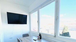 Moderni huone, jossa on suuri ikkuna, seinälle asennettu taulutelevisio, puinen pöytä ja neljä tuolia sekä patteri ikkunan alla.