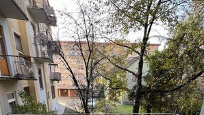 En balkon med flisegulv, gelænder i metal og udsigt til omkringliggende bygninger og træer.