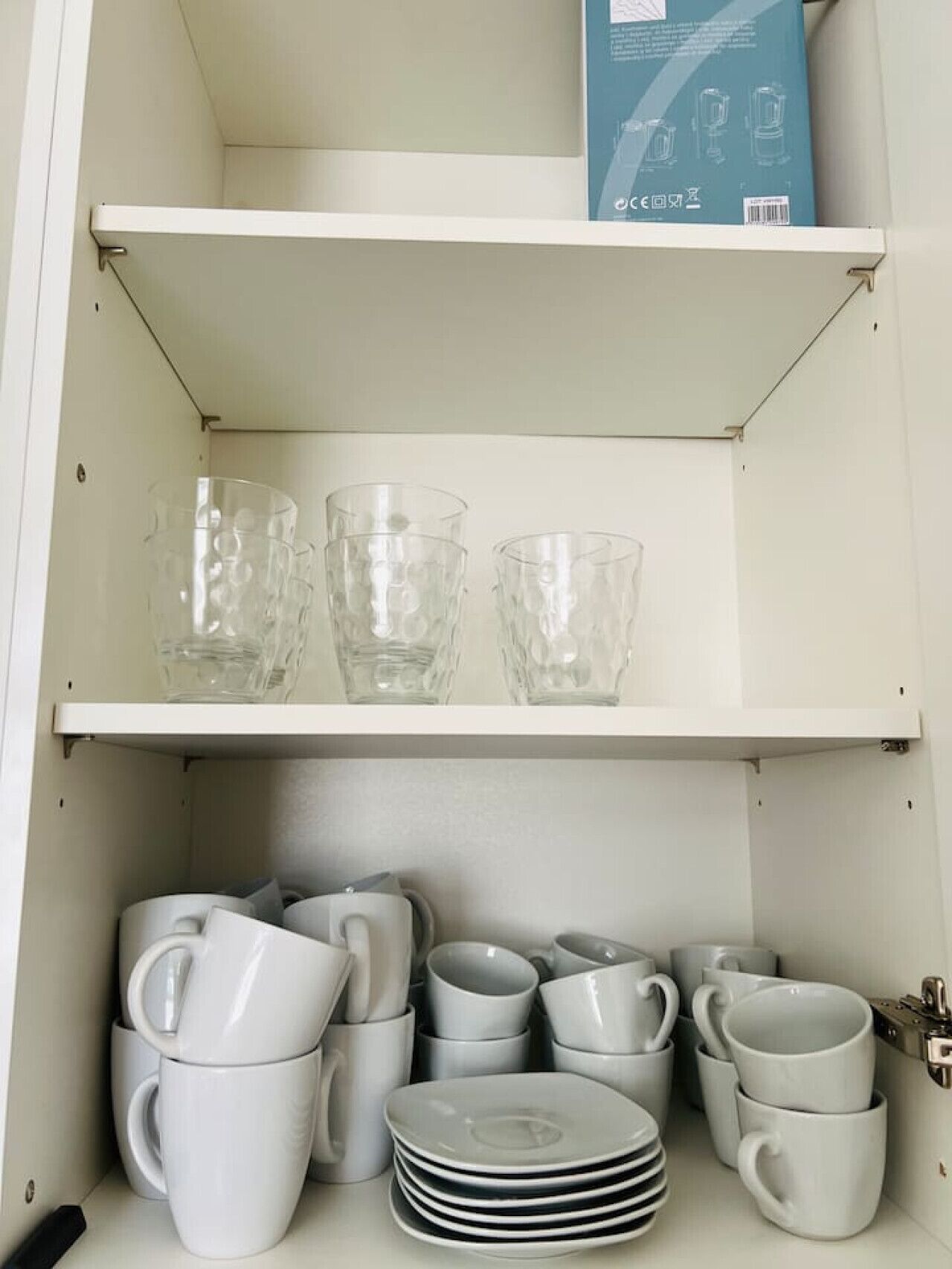 A kitchen cabinet with two shelves; the top shelf holds three clear glass cups, and the bottom shelf contains stacked white mugs and plates.