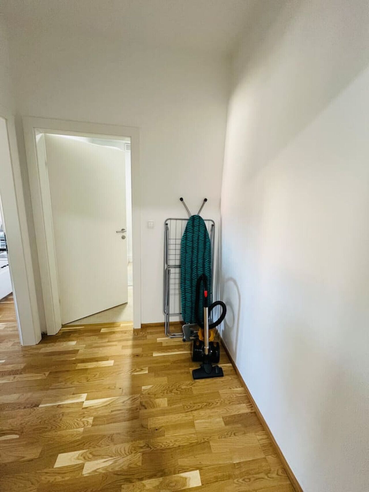 Een gang met houten vloer, een metalen droogrek met kleding en een stofzuiger.