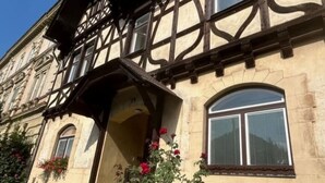 A traditional half-timbered building with a prominent entrance, a small garden with red flowers, and a clear blue sky.