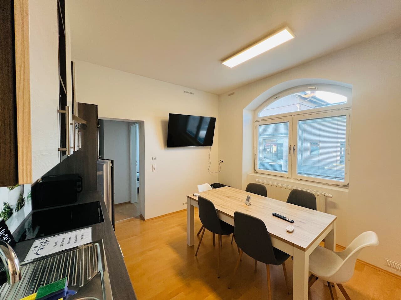 Una cocina moderna con estufa integrada, un comedor con mesa y sillas, un televisor de pantalla plana en la pared y una ventana con persianas.