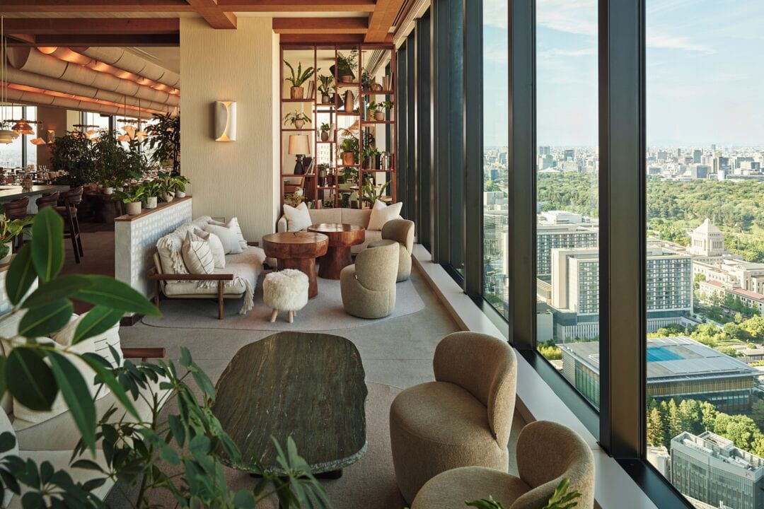 A high-rise interior with a city view, a seating area with sofas and a coffee table, and a bookshelf with plants.