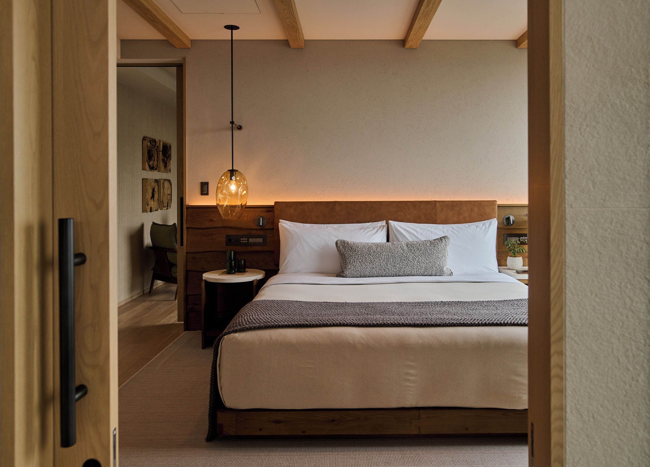 Un dormitorio con cabecera de madera, cama con ropa de cama blanca, mesita de noche redonda y lámpara colgante.