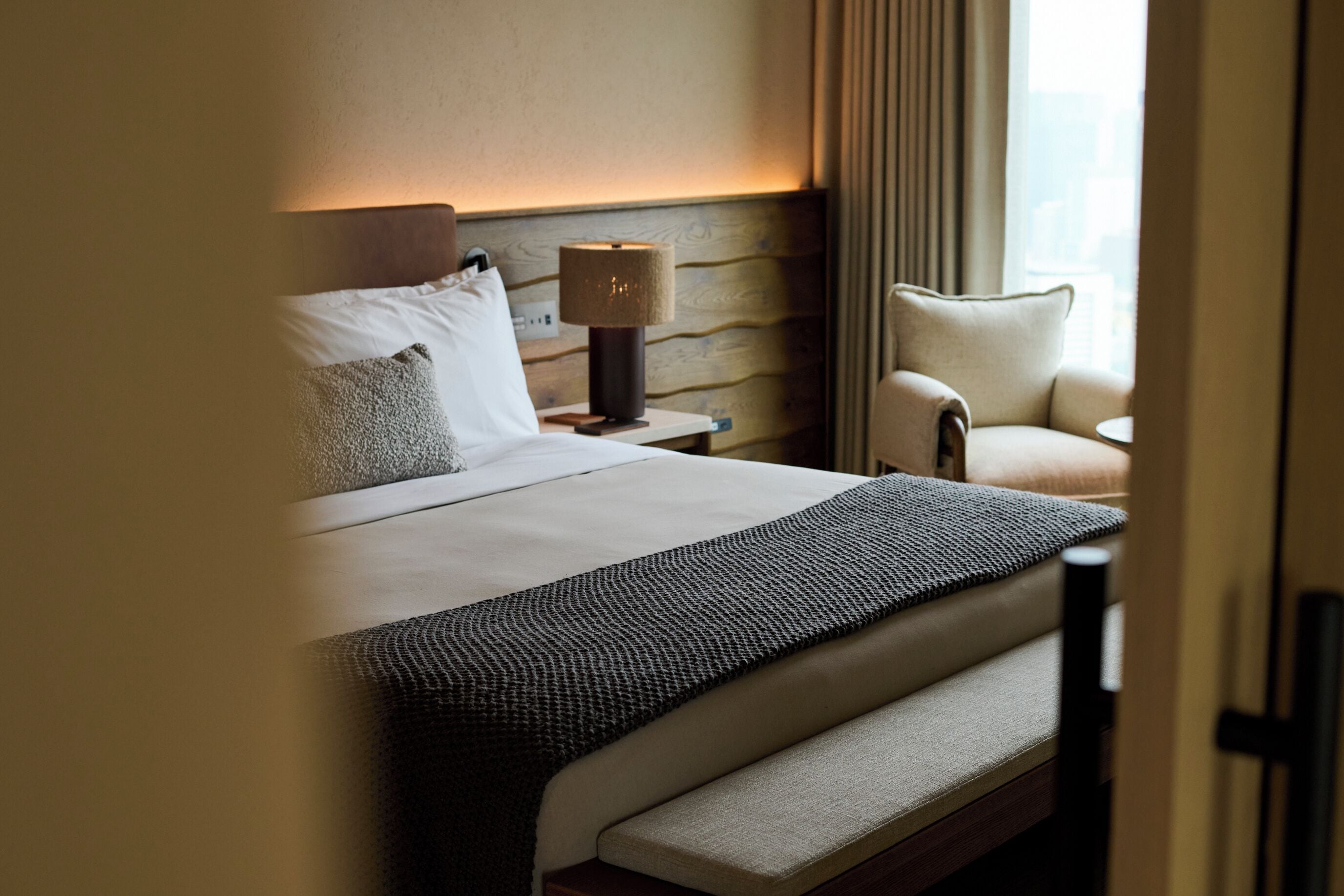 A hotel room with a bed, bedside lamp, chair, and a view of the city.