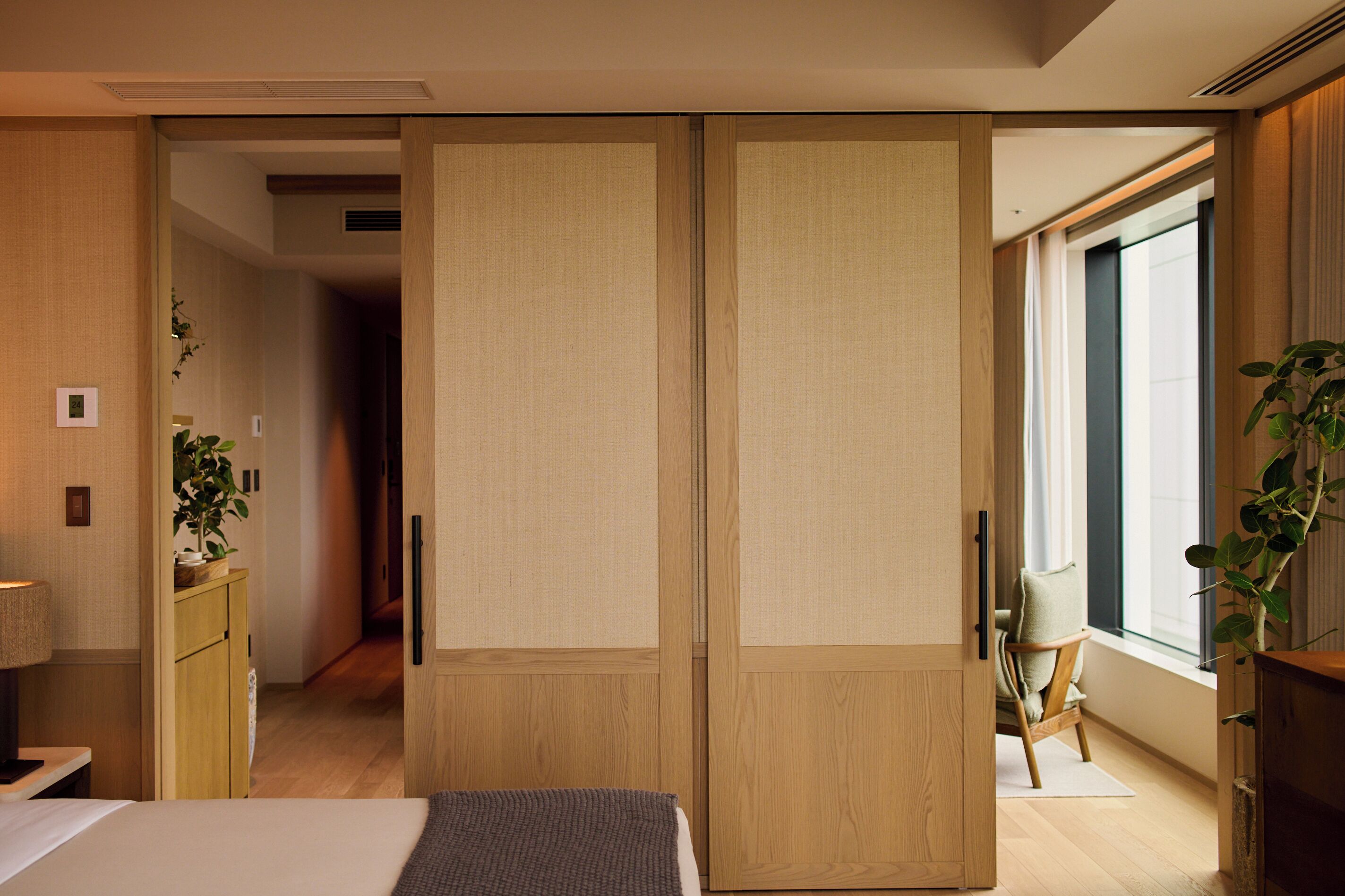 A hotel room with a bed, a desk, a chair, a sliding door, and a window with a plant.