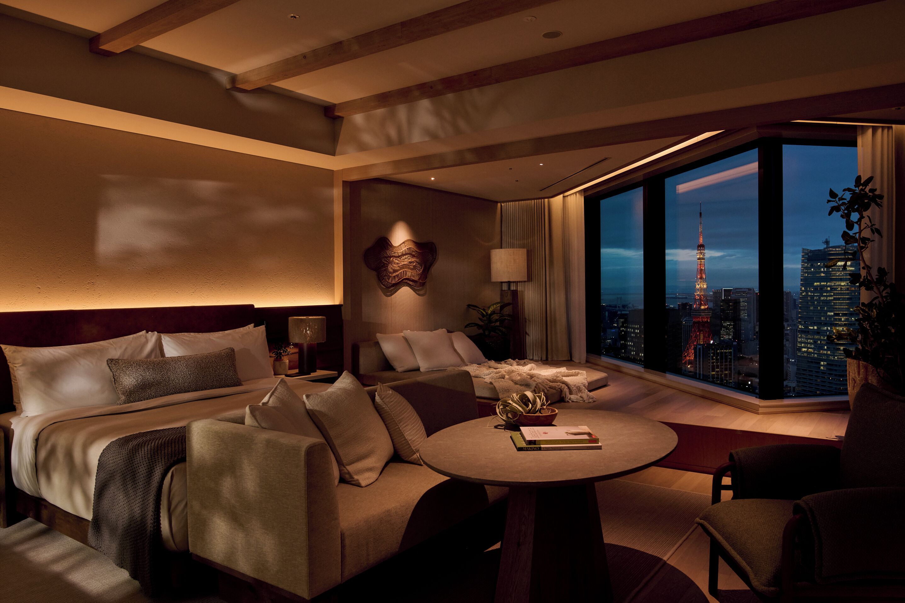 A modern hotel room with a large bed, a sofa, a small table, and a view of a cityscape at night.