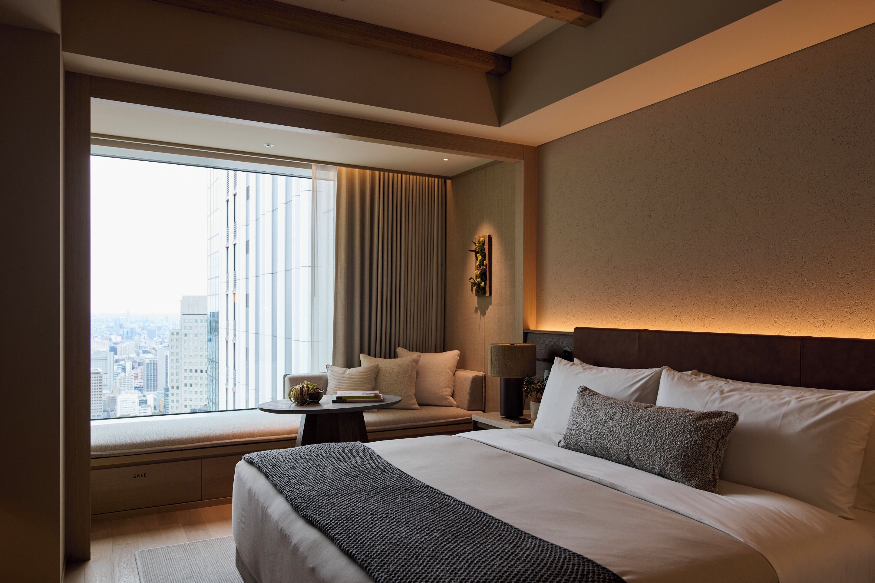 Ein modernes Hotelzimmer mit einem großen Bett, einer Sitzecke mit Tisch und Blick auf die Stadt durch große Fenster. Draußen ist eine Stadtansicht sichtbar.
