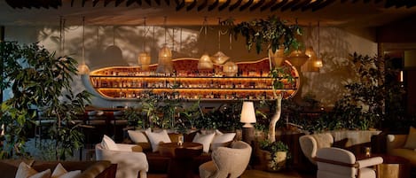 Una zona de bar con estanterías llenas de botellas, luces colgantes y asientos decorados con plantas.