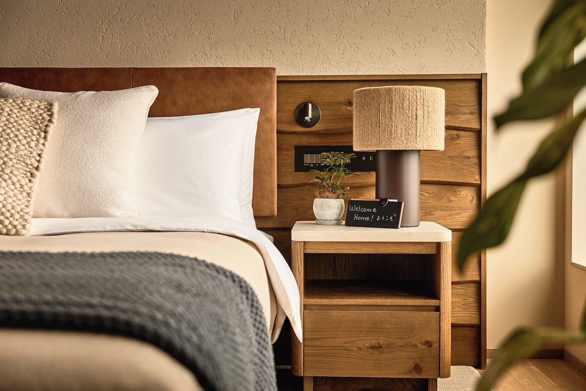 Un lit bien fait, avec une tête de lit en bois, une lampe de chevet, une petite plante et un panneau d’accueil posé sur une table de chevet en bois.