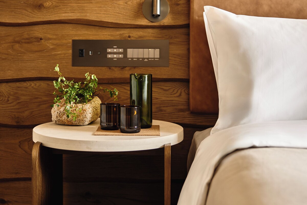 Una habitación de hotel moderna con un cabecero de madera, una mesita de noche con una planta en maceta, un control remoto y una cama con ropa de cama blanca.