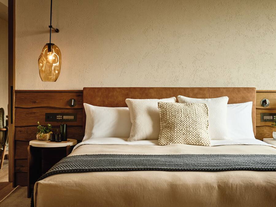 Une chambre d’hôtel avec une tête de lit en bois, un grand lit avec une literie blanche, un plaid texturé, une petite table de chevet avec une plante et un luminaire suspendu.