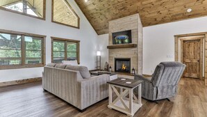 A spacious living room with a fireplace, wooden ceiling, and large windows. - Mountaintown Chalet | Ellijay, GA (Ellijay)