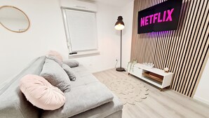 Una sala de estar moderna con un sofá gris, una consola blanca, un letrero de Netflix, una lámpara de pie y una ventana con persianas.
