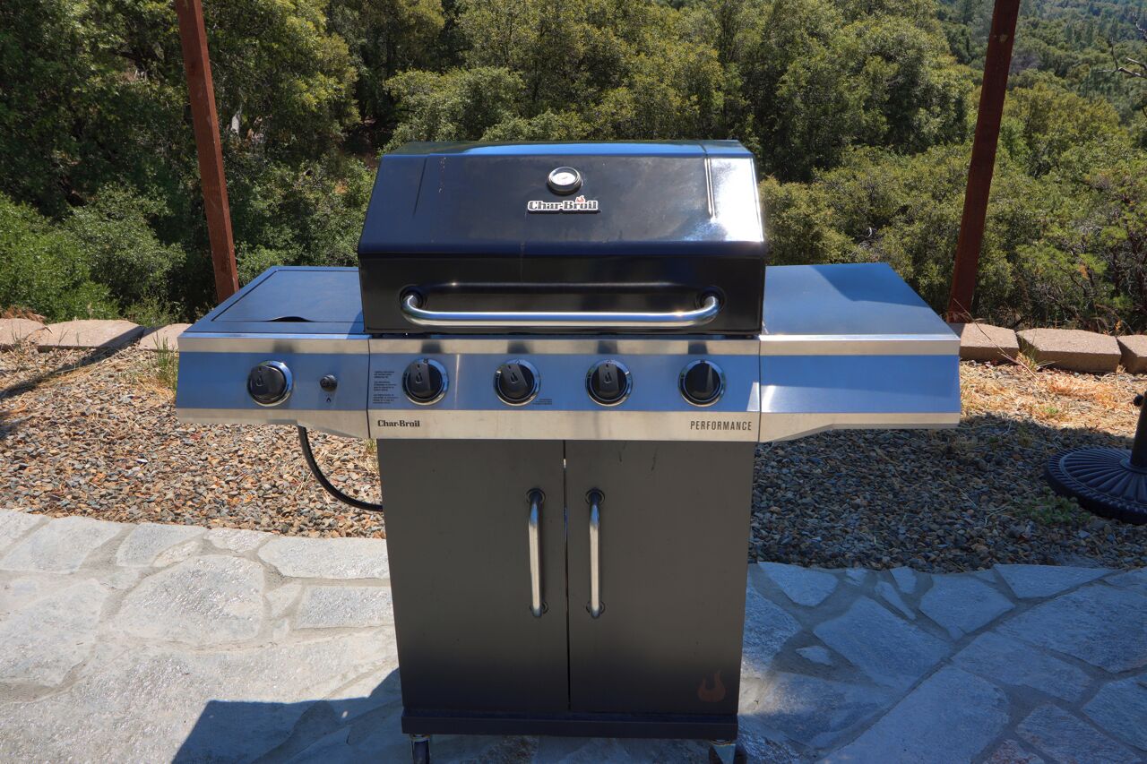 A Char-Broil Performance grill with four burners, a digital display, and a stainless steel finish, set on a stone patio with a forest backdrop.