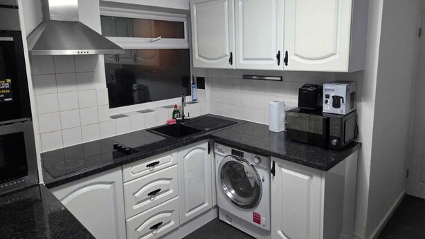 A compact kitchen with white cabinets, a stainless steel range hood, a microwave, a kettle, and a washing machine.