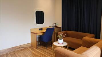 A compact hotel room with a wooden desk, a blue chair, a brown sofa, a small round table with a potted plant, and a window with a black curtain.
