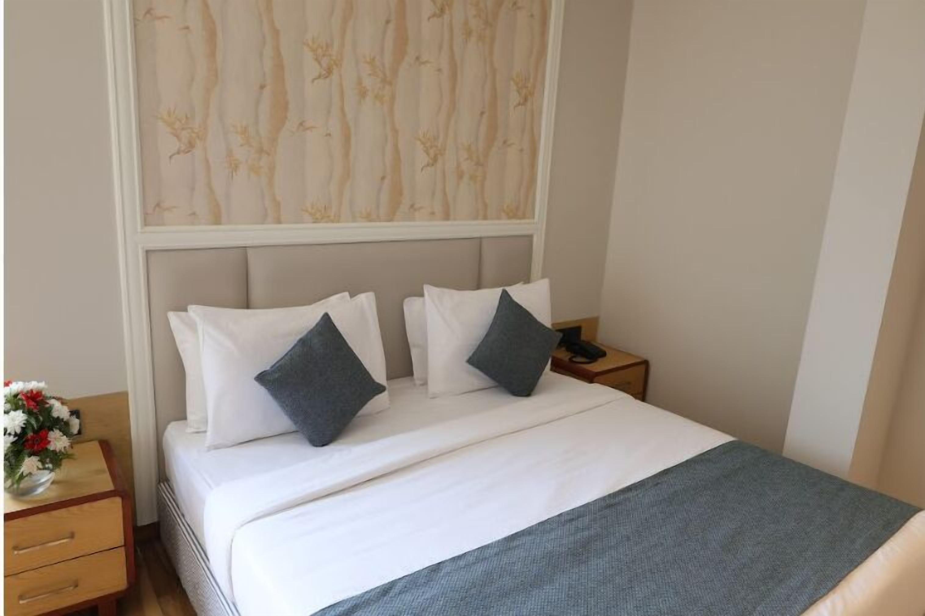 A neatly made bed with white linens and two pillows, a bedside table with a vase of flowers, and a wall with a patterned wallpaper.