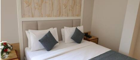 A neatly made bed with white linens and two pillows, a bedside table with a vase of flowers, and a wall with a patterned wallpaper.