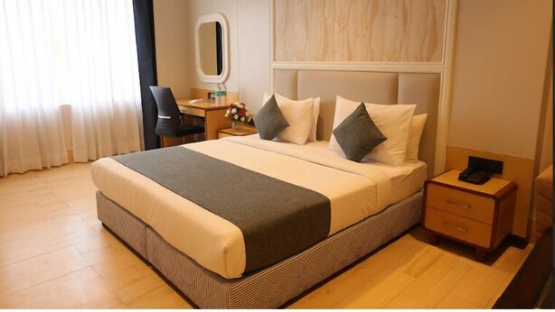 A hotel room with a large bed, a bedside table, a chair, a desk, a mirror, and a window with curtains. - Prakash Continental (Bareilly)