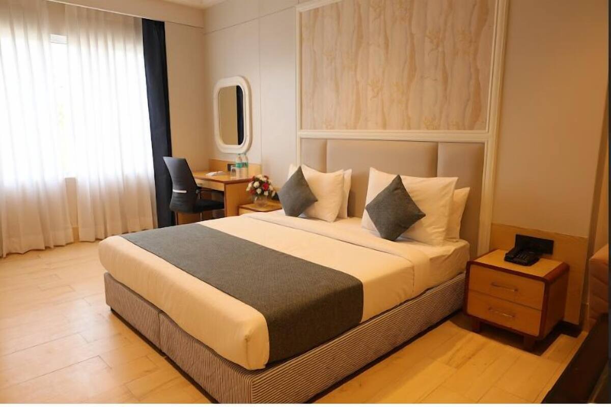 A hotel room with a large bed, a bedside table, a chair, a desk, a mirror, and a window with curtains.