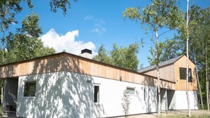 Una casa moderna con fachada blanca y techo de madera, rodeada de árboles y cielo despejado.