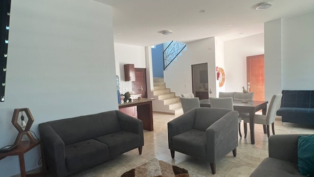 Amplio salón con diseño moderno, que incluye un gran sofá, una mesa de centro y una alfombra estampada. Una escalera conduce a un nivel superior, y la habitación tiene una paleta de colores neutros.