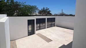 Un patio en la azotea con un muro blanco, un portón negro y un cielo azul despejado.