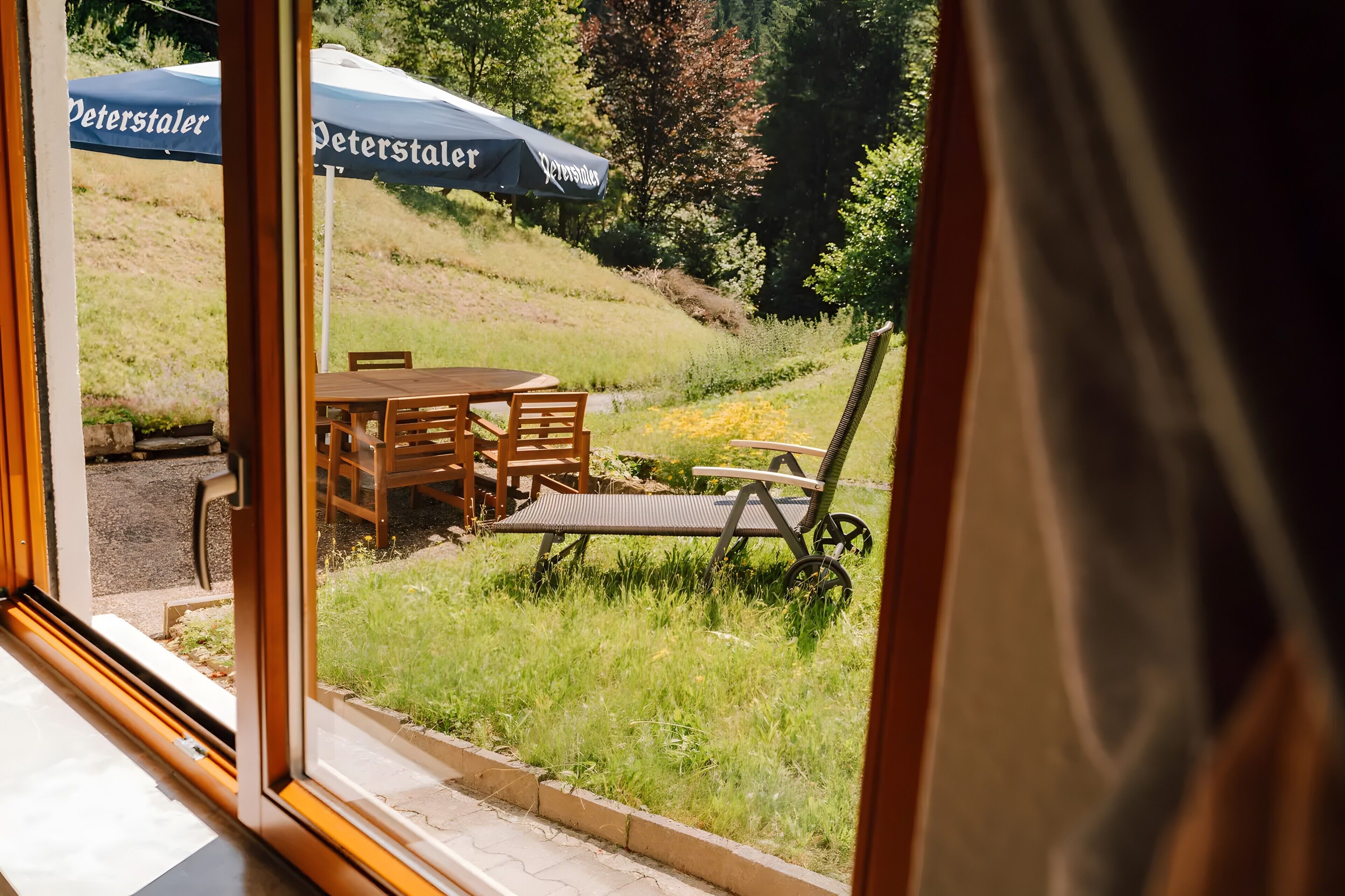 Blick durch eine Glastür auf einen Essbereich mit Außenplätzen, darunter ein Tisch und Stühle, sowie zwei Peterstaler Sonnenschirme im Hintergrund.