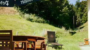 Outdoor seating with a table and chairs, a blue umbrella with text, and a grassy area with trees. - Bästenbach Apartment: Idyllic Quiet Location with Mountain Views, Private Terrace & Wi-Fi (Bad Peterstal-Griesbach)
