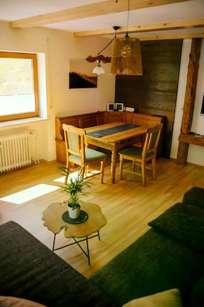 A cozy living room with a wooden dining table, chairs, a green sofa, a small side table with a plant, and a large window.