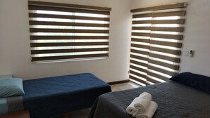 Quarto com duas camas, cada uma com colcha azul acolchoada. Há duas janelas com persianas horizontais, e uma tomada visível à direita das janelas.