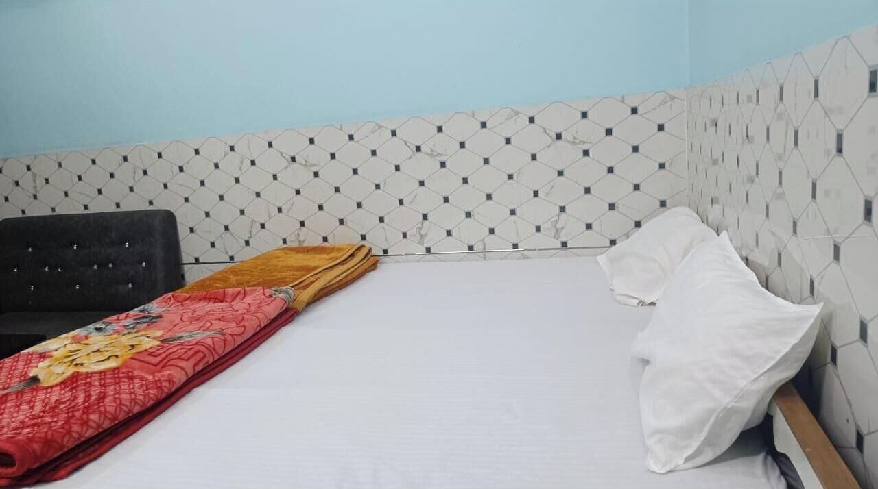 A single bed with a white mattress and a red blanket, a black upholstered chair, and a white pillow on the right side.