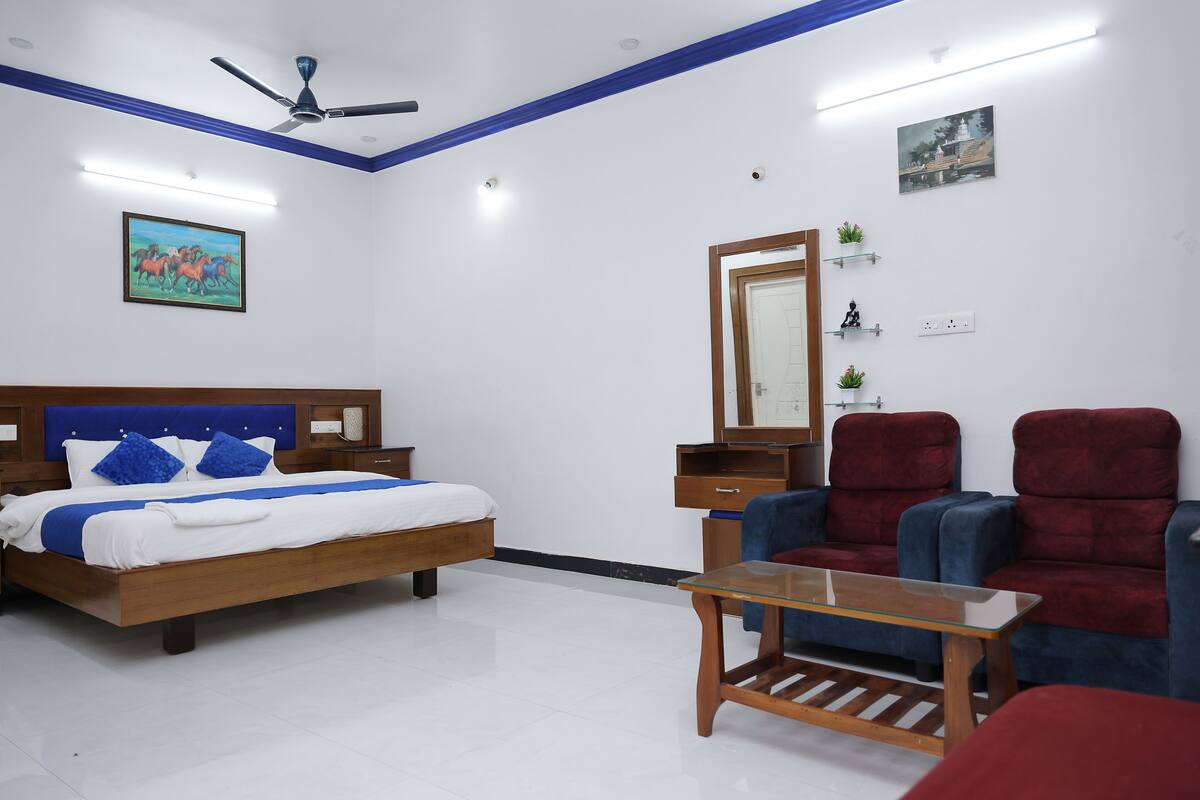 A bedroom with a bed, a desk, two chairs, a ceiling fan, and a painting on the wall.