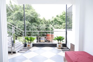 Un balcón con suelo a cuadros, dos plantas en macetas, una cama roja y vistas a árboles y un camión.
