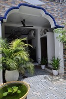 Un patio con suelo de baldosas, una planta grande en maceta, un pequeño estanque con lirios y un ventilador de techo.