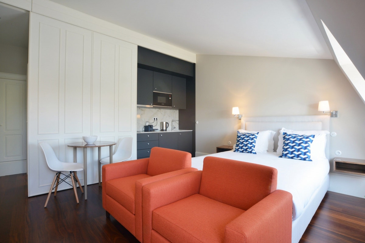 Una habitación de hotel moderna con una cama, sofá naranja, sillas blancas, una mesita pequeña y una kitchenette.