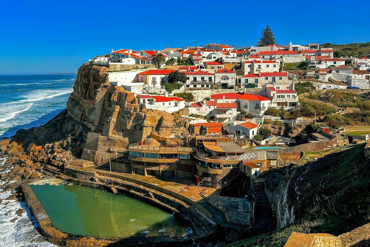 Un pueblo costero con edificios sobre un acantilado, un muro marítimo y una pequeña piscina.