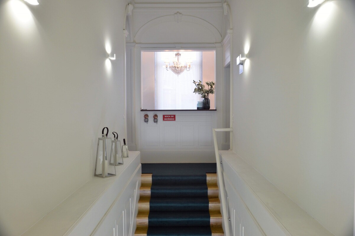 Una escalera con alfombra azul que lleva a una habitación con candelabro, un cartel que indica una chimenea y luces fijadas en la pared.